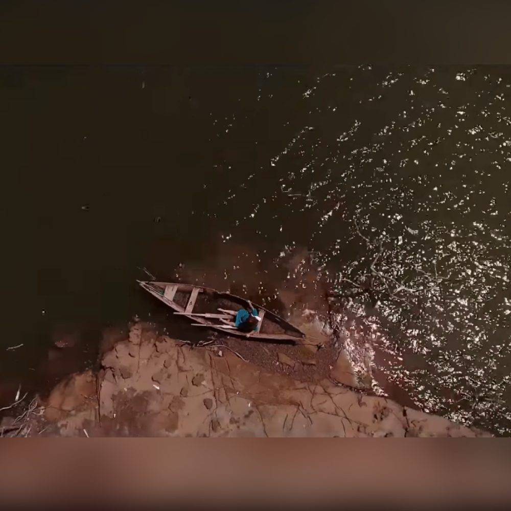 Cena do filme O Pescador e o Rio, baseado na lenda do Cabeça de Cuia. Imagem vista de cima. Mostra uma mulher com um vestido azul sentada em uma canoa nas margens de um rio.