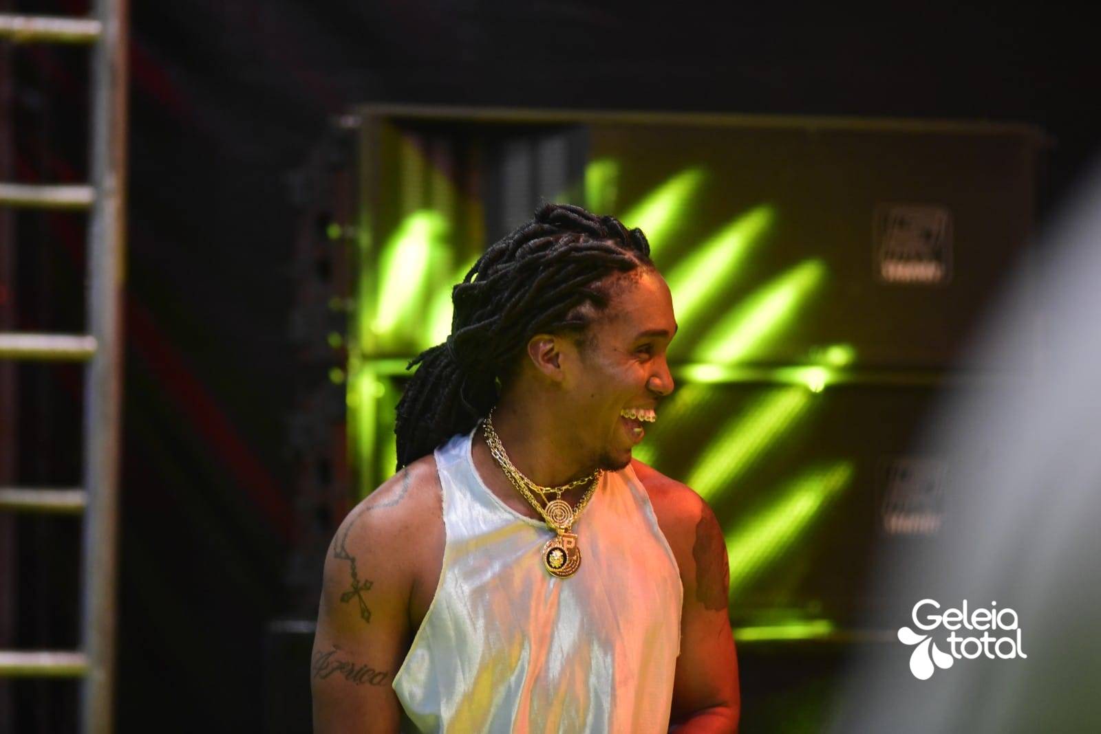 Foto do rapper Preto Kedé sorrindo e em cima do palco. “deixa a chuva cair / deixa o céu se abrir / deixa o mundo girar / não só em torno de ti”.