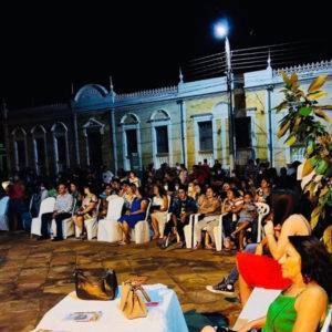 Foto da praça à noite cheia de pessoas sentadas assistindo às apresentações.