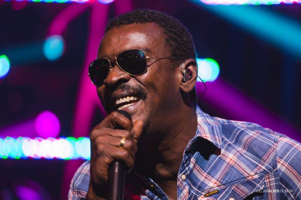 Foto de Seu Jorge, cantando com microfone, ele usa óculos escuros.